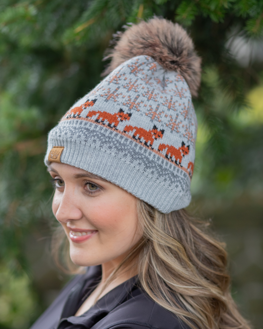 Woman wearing a gray knit hat with fox pattern and brown pom-pom against a blurred green background.