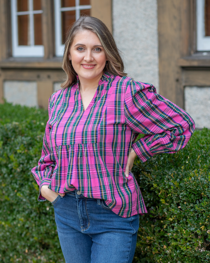 Woman wearing a pink plaid shirt standing outdoors with greenery and a building in the background.