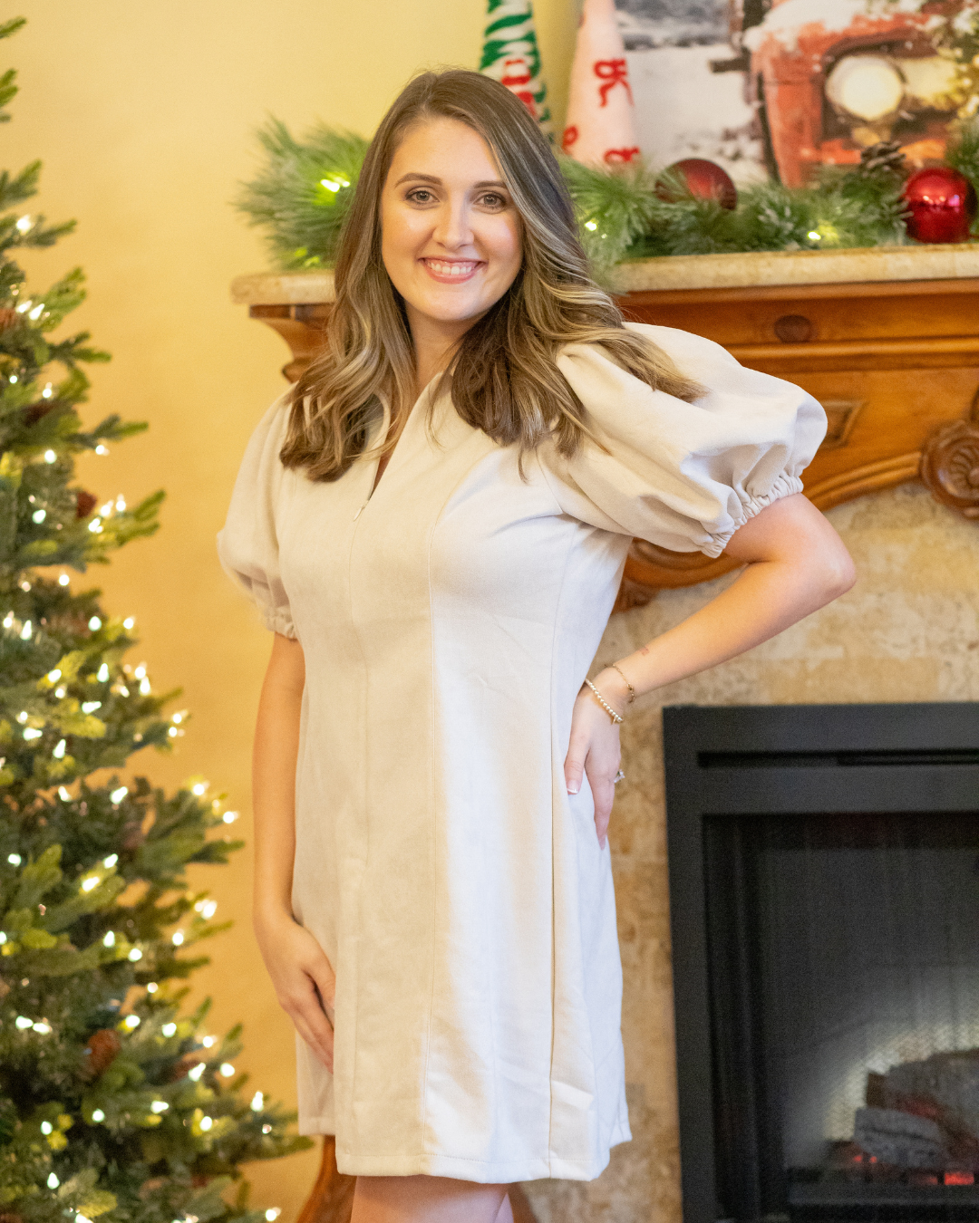 Woman in a tan dress standing in front of a decorated Christmas tree and fireplace.