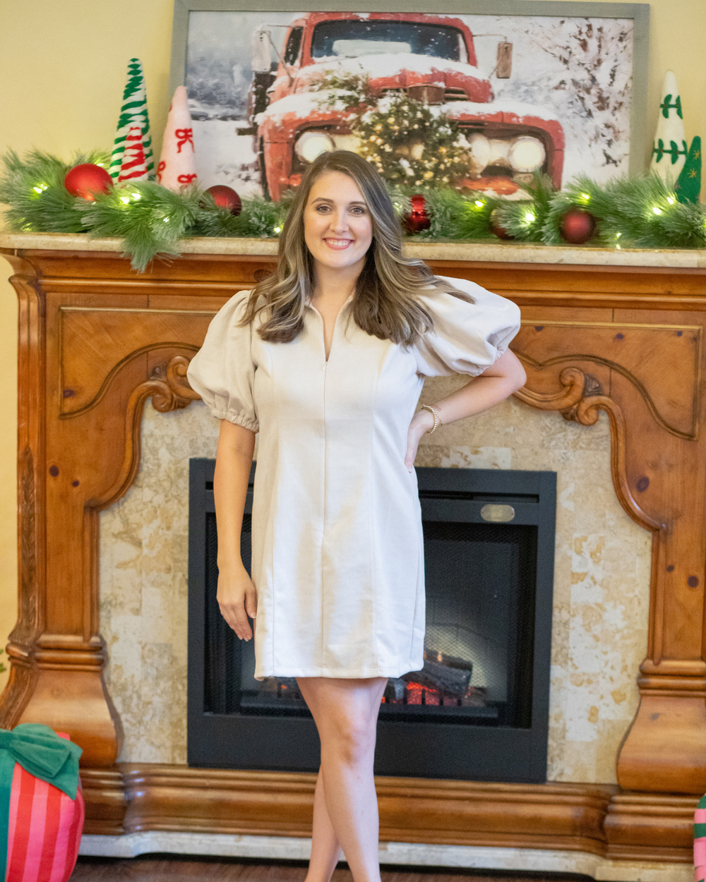 Woman in a tan dress standing in front of a decorated fireplace with Christmas decorations.