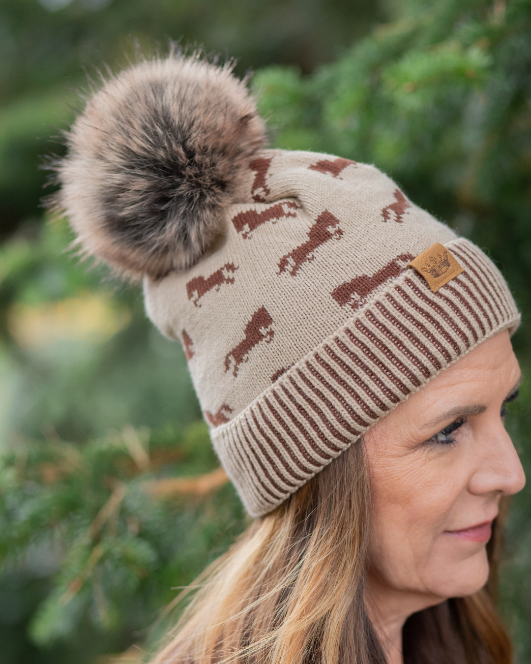 Beige knit beanie with brown horse pattern and fur pom-pom worn by a person against a blurred green background.