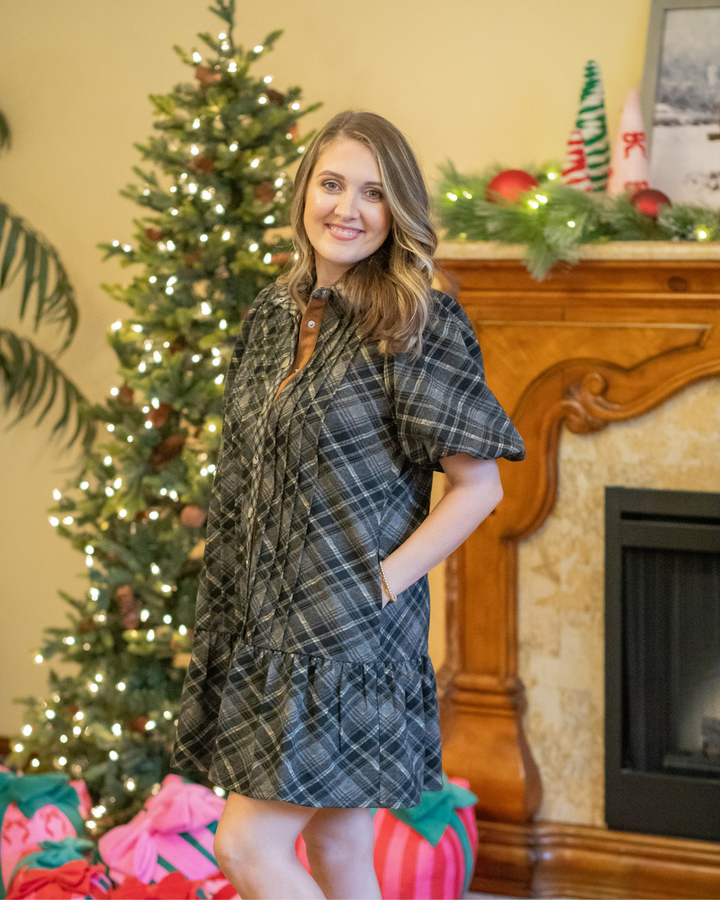 Woman in a plaid dress standing in front of a Christmas tree and fireplace.