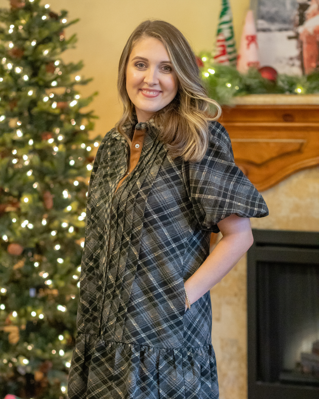 Woman wearing a plaid dress in front of a decorated Christmas tree and fireplace.