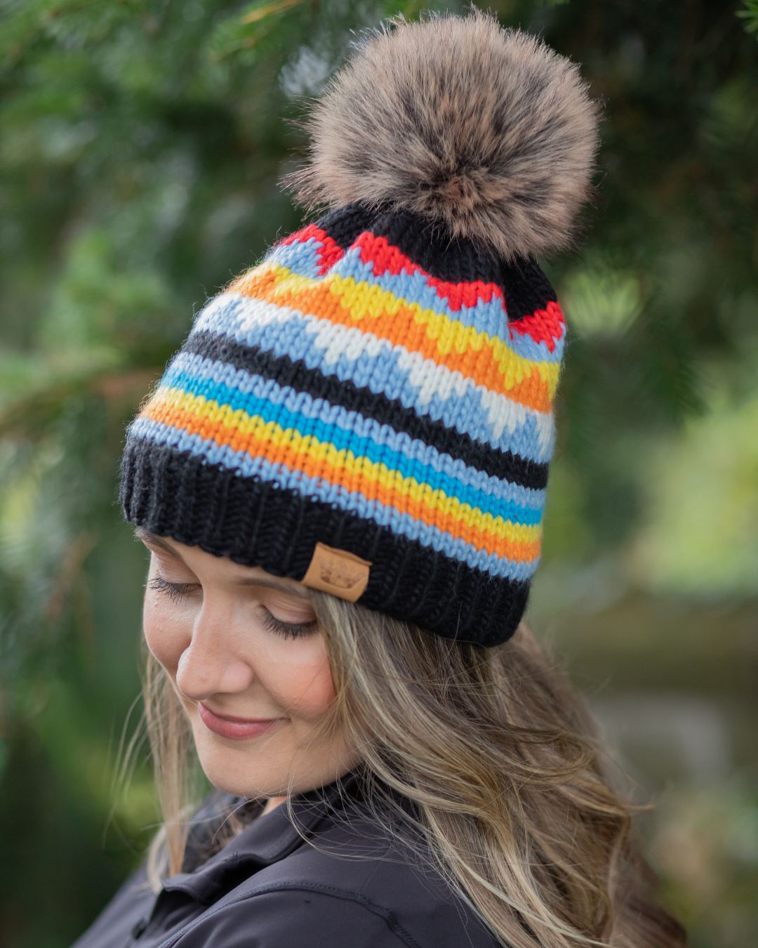 Person wearing a colorful striped beanie with a pom-pom in front of green foliage.