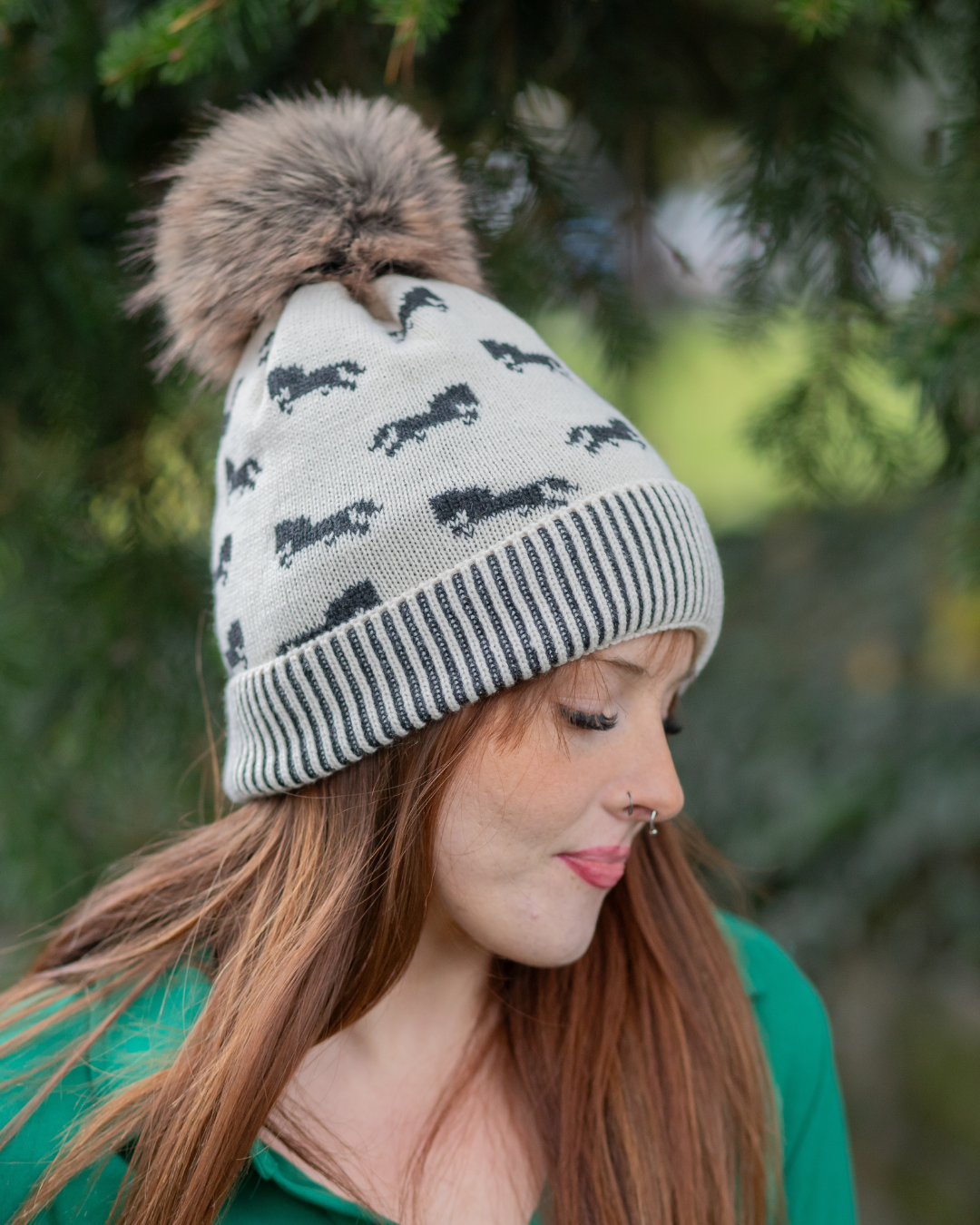 Woman wearing a knitted hat with horse patterns and a fur pom-pom in a forest setting.