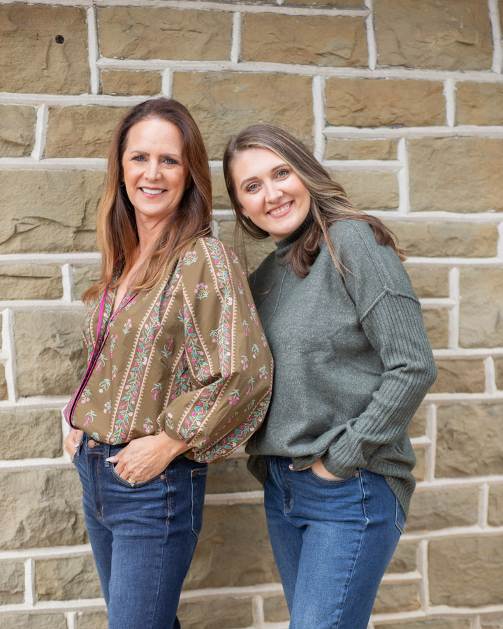 Two women standing against a brick wall, wearing patterned tops and jeans.