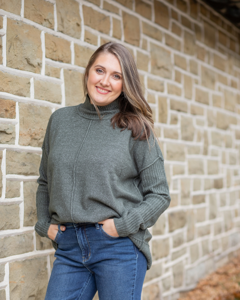Woman wearing a gray sweater and blue jeans standing against a brick wall.