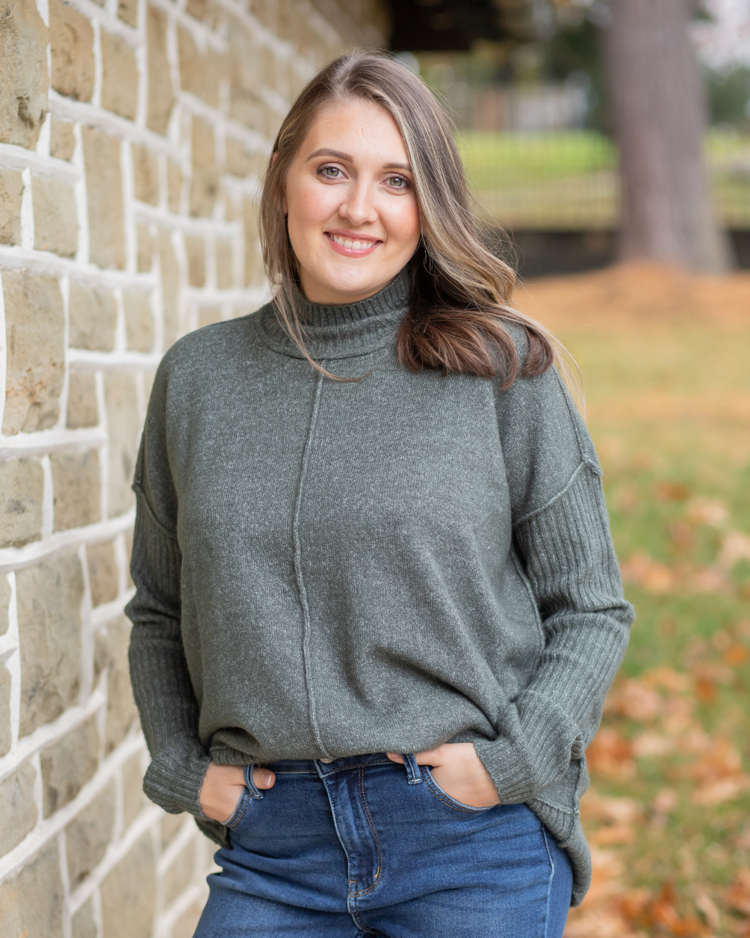 Woman wearing a green sweater and blue jeans standing against a brick wall.