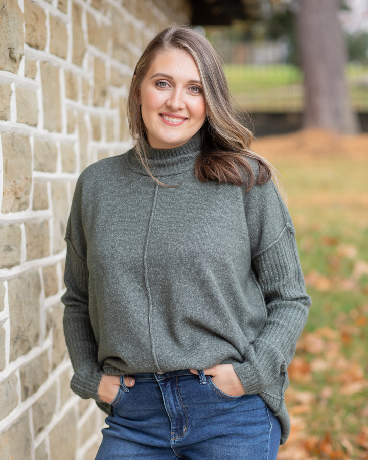 Woman wearing a green sweater and blue jeans standing against a brick wall.