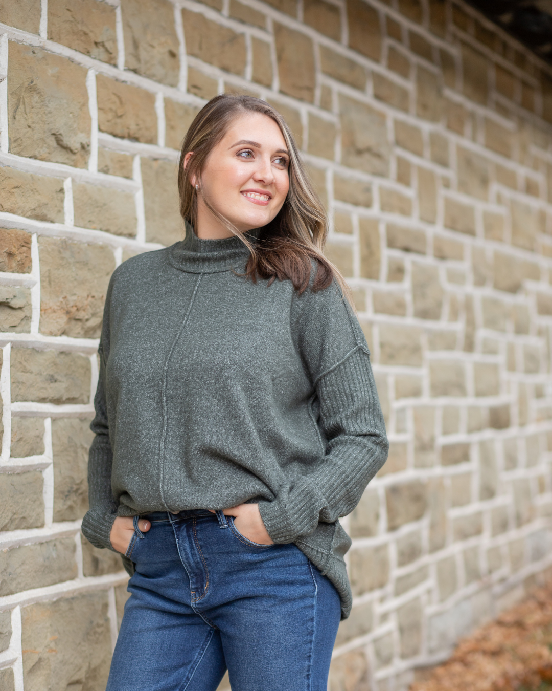 Woman wearing a green sweater and blue jeans standing against a brick wall.