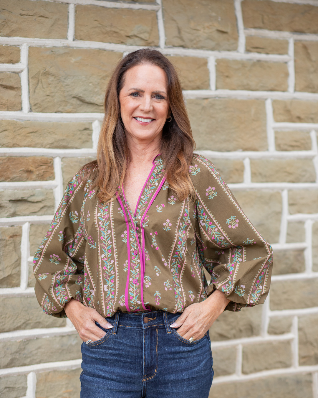 Woman wearing a patterned blouse and jeans standing against a brick wall.