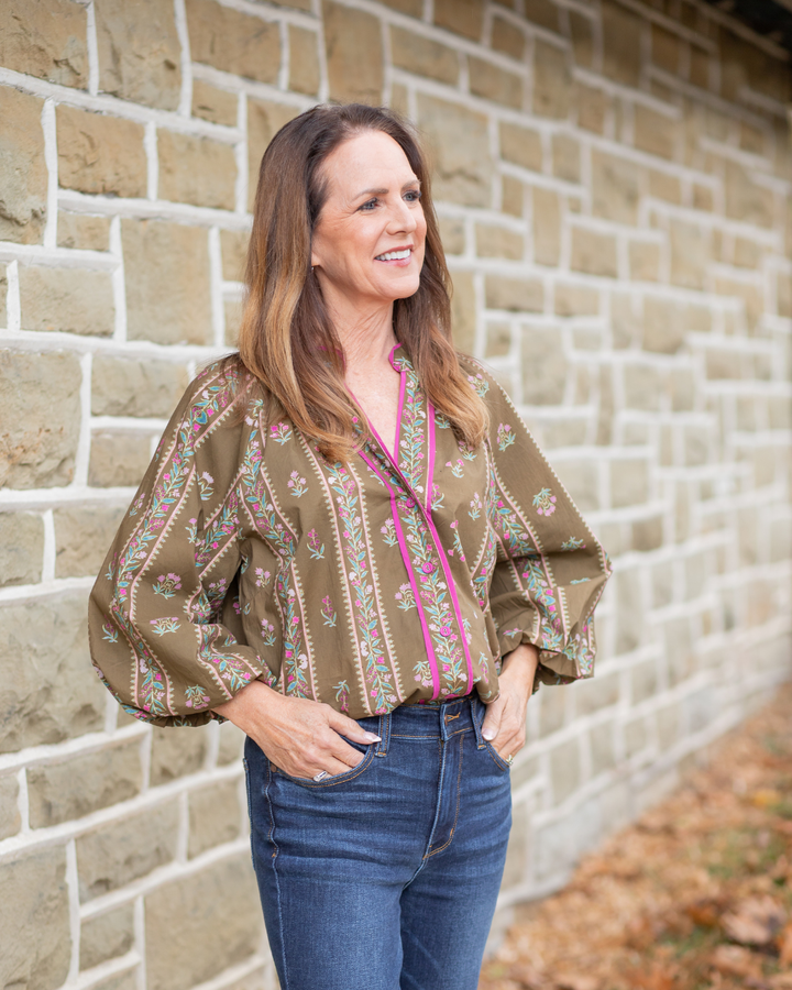 Woman wearing a patterned blouse and jeans standing against a brick wall.