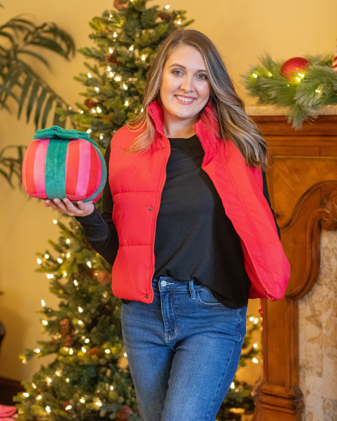 Woman in a red vest holding a striped gift box in front of a decorated Christmas tree.