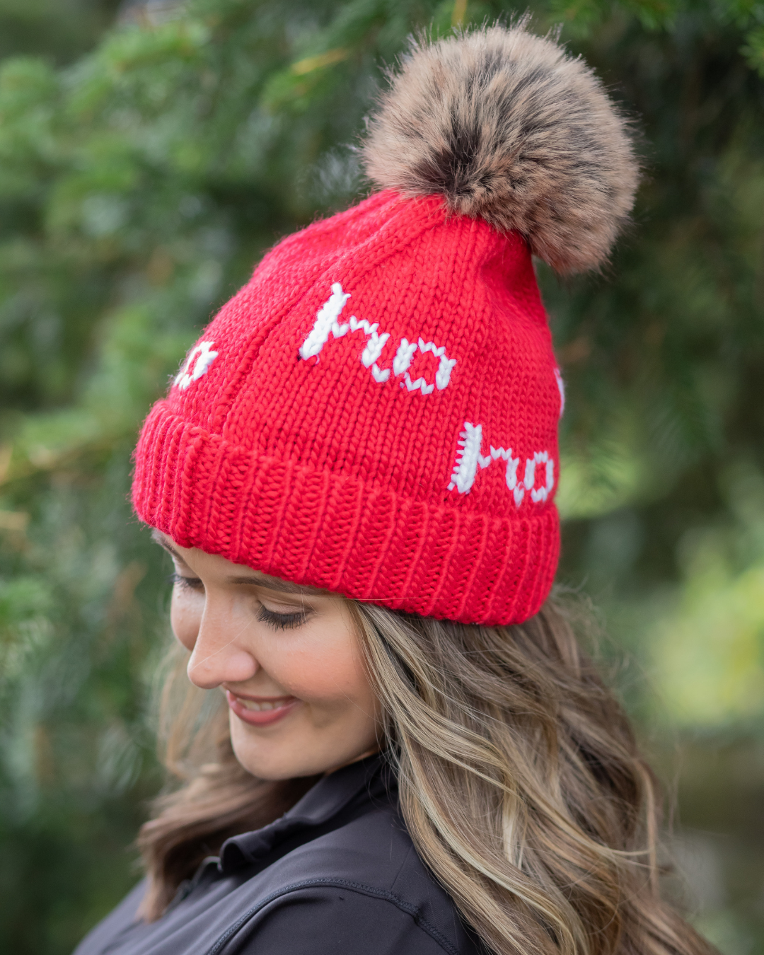 Person wearing a red knit hat with a fur pom-pom and 'ho ho ho' text, standing against a blurred green background.